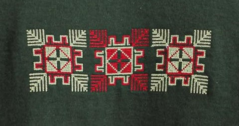 A green t-shirt with a central embroidered Tatreez design, displayed on a hanger against a neutral background with a plant to the side.
