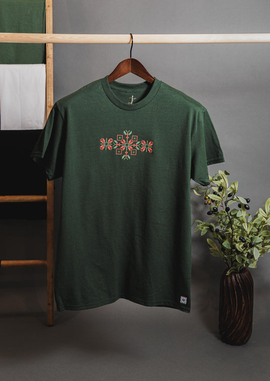 A green t-shirt with a central embroidered Tatreez design, displayed on a hanger against a neutral background with a plant to the side.