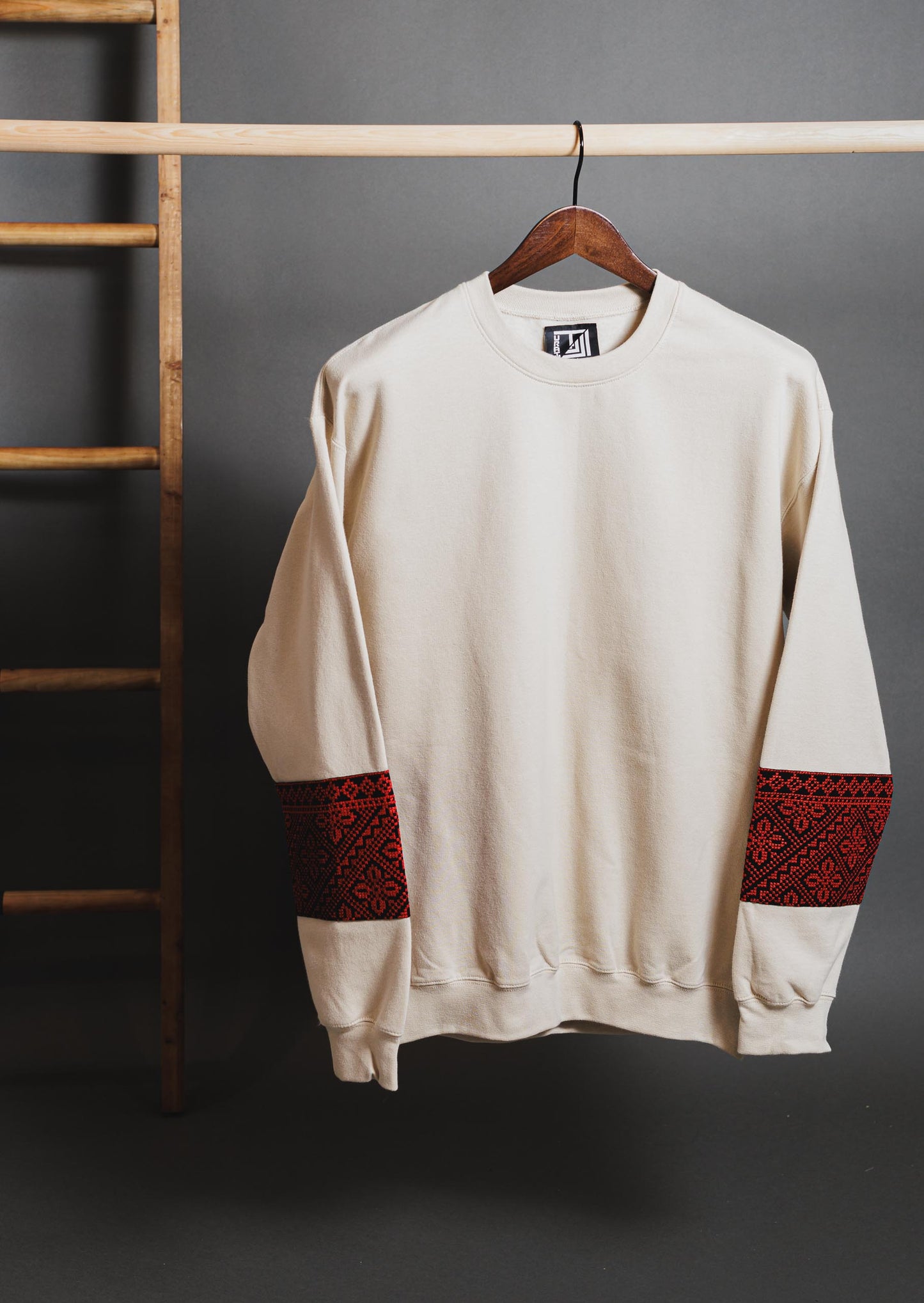 A navy sweatshirt with red Tatreez bracelet patterns on the sleeves, displayed on a hanger against a gray background.