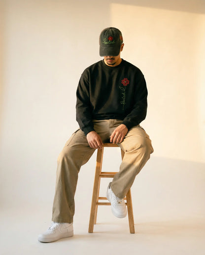 Person sitting on a stool wearing a black sweatshirt with a red rose design, beige pants, and white sneakers.
