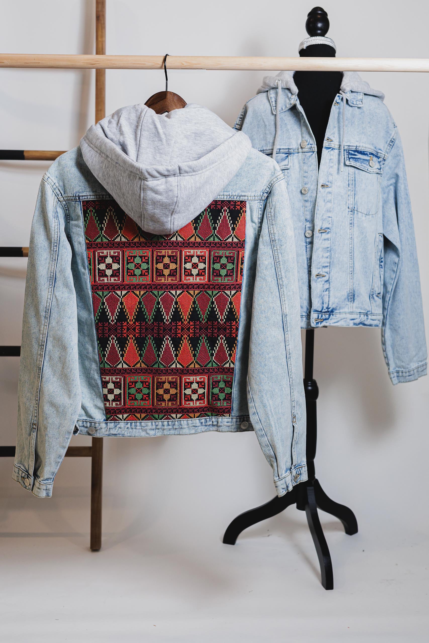 Unisex light blue denim jacket with gray hood and vibrant Palestinian Tatreez embroidery on back.